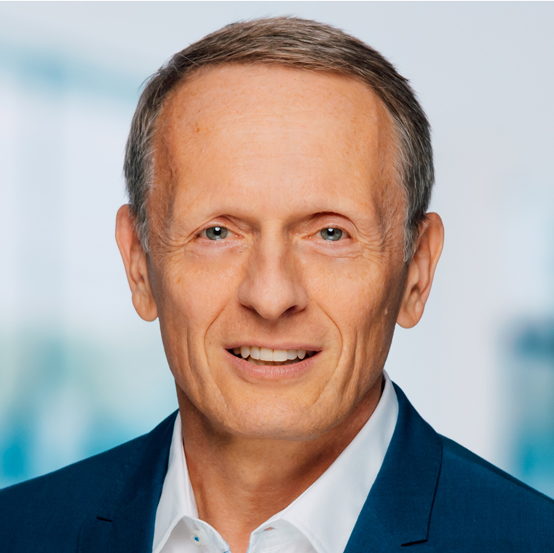 Ein Mann mittleren Alters mit kurzem hellbraunem Haar, einem blauen Jackett und einem weißen Hemd, posiert vor einem unscharfen hellblauen Hintergrund.